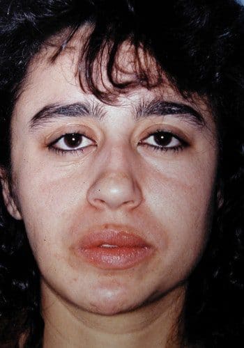 A close-up image of a woman with dark curly hair, neutral facial expression, prominent eyebrows, and visible lipstick.