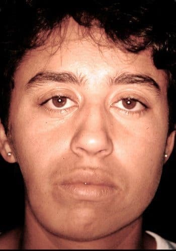 Close-up of a person with short dark hair, neutral expression, and earrings, looking directly at the camera against a dark background.