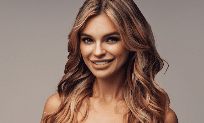 Smiling woman with long, wavy blonde hair, wearing a strapless top, poses in front of a plain background.