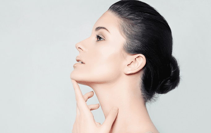 Profile view of a person with dark hair in a bun, touching their chin and looking upwards against a plain background.