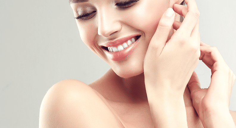 A woman with a radiant smile touches her ear, displaying smooth skin and closed eyes.