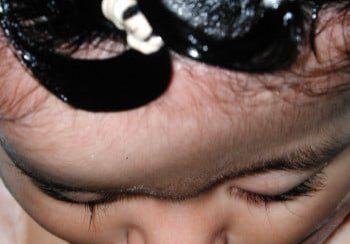 A child with their head shaved and wearing a black wig.