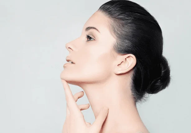 Profile view of a person with dark hair in a bun, touching their chin and looking upwards against a plain background.