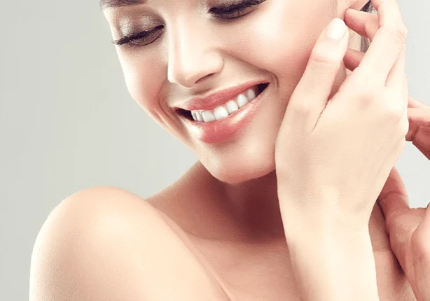 A woman with a radiant smile touches her ear, displaying smooth skin and closed eyes.